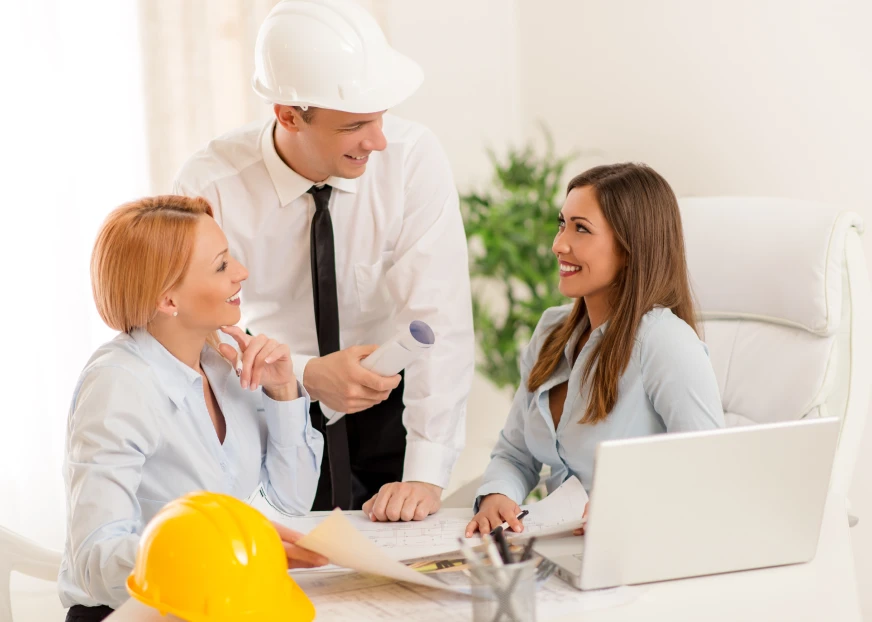 Three professionals discussing documents at a desk, with a laptop and a yellow hard hat visible in the foreground.