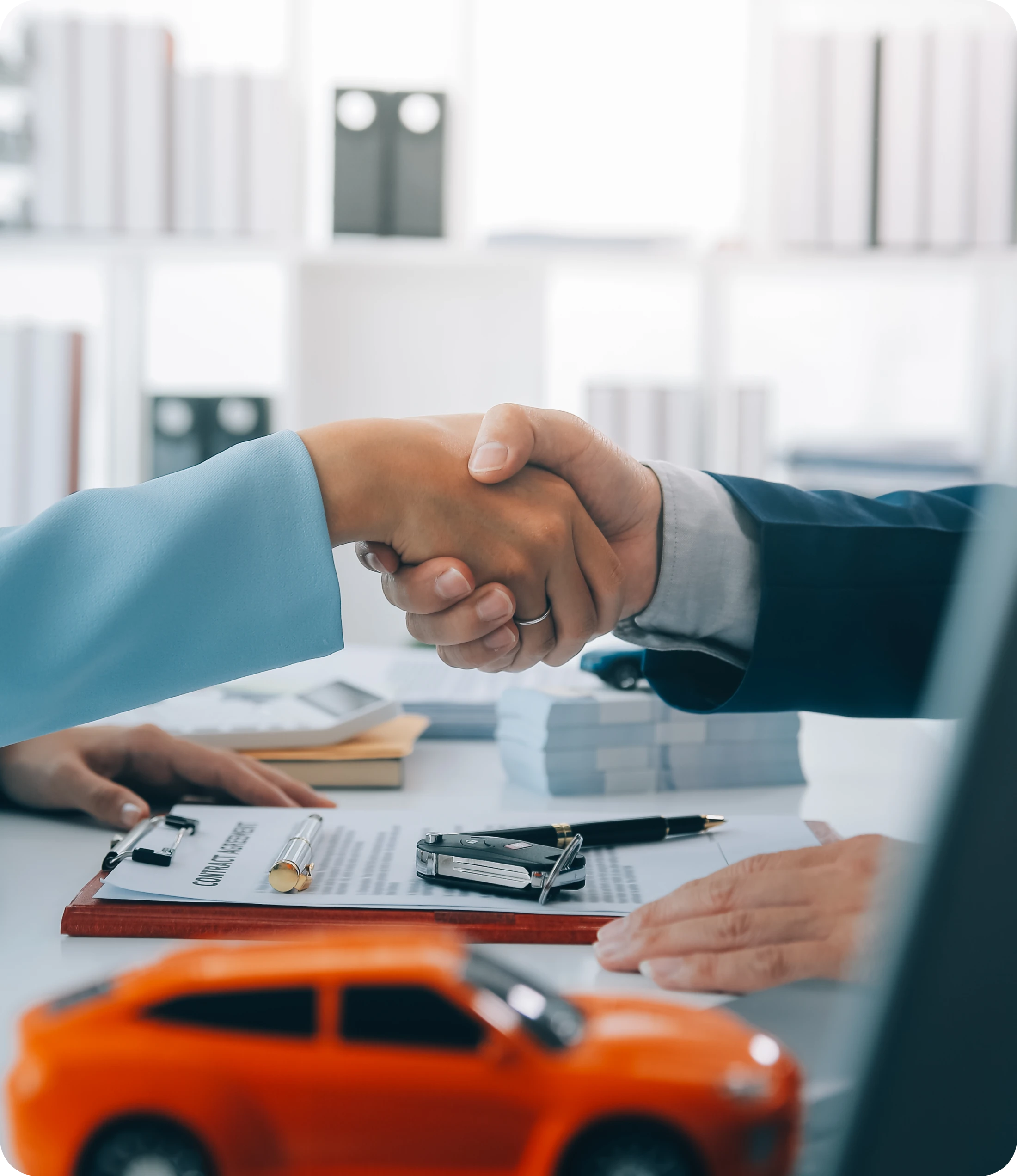 Handshake Over Desk With Toy Orange Car, Car Keys, & Insurance Document