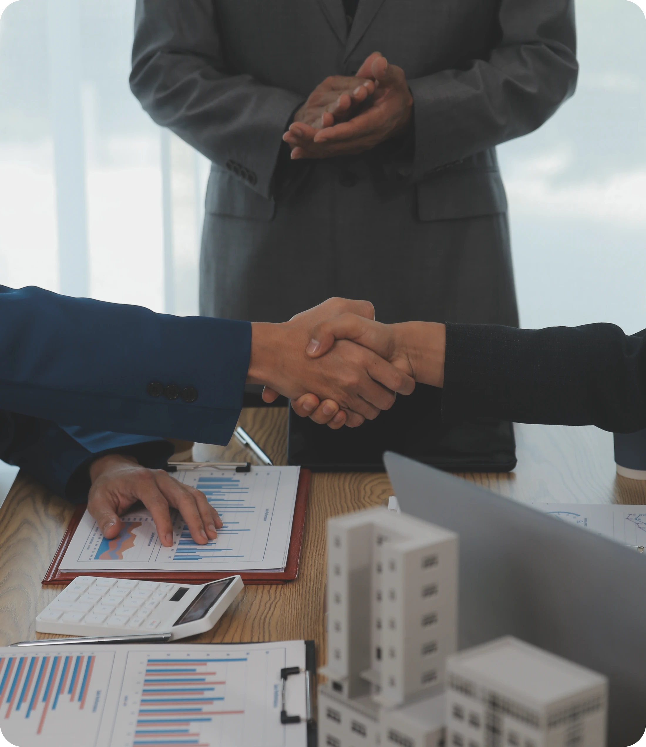 Two People Shaking Hands Over Documents & A Suited Figure Standing Behind