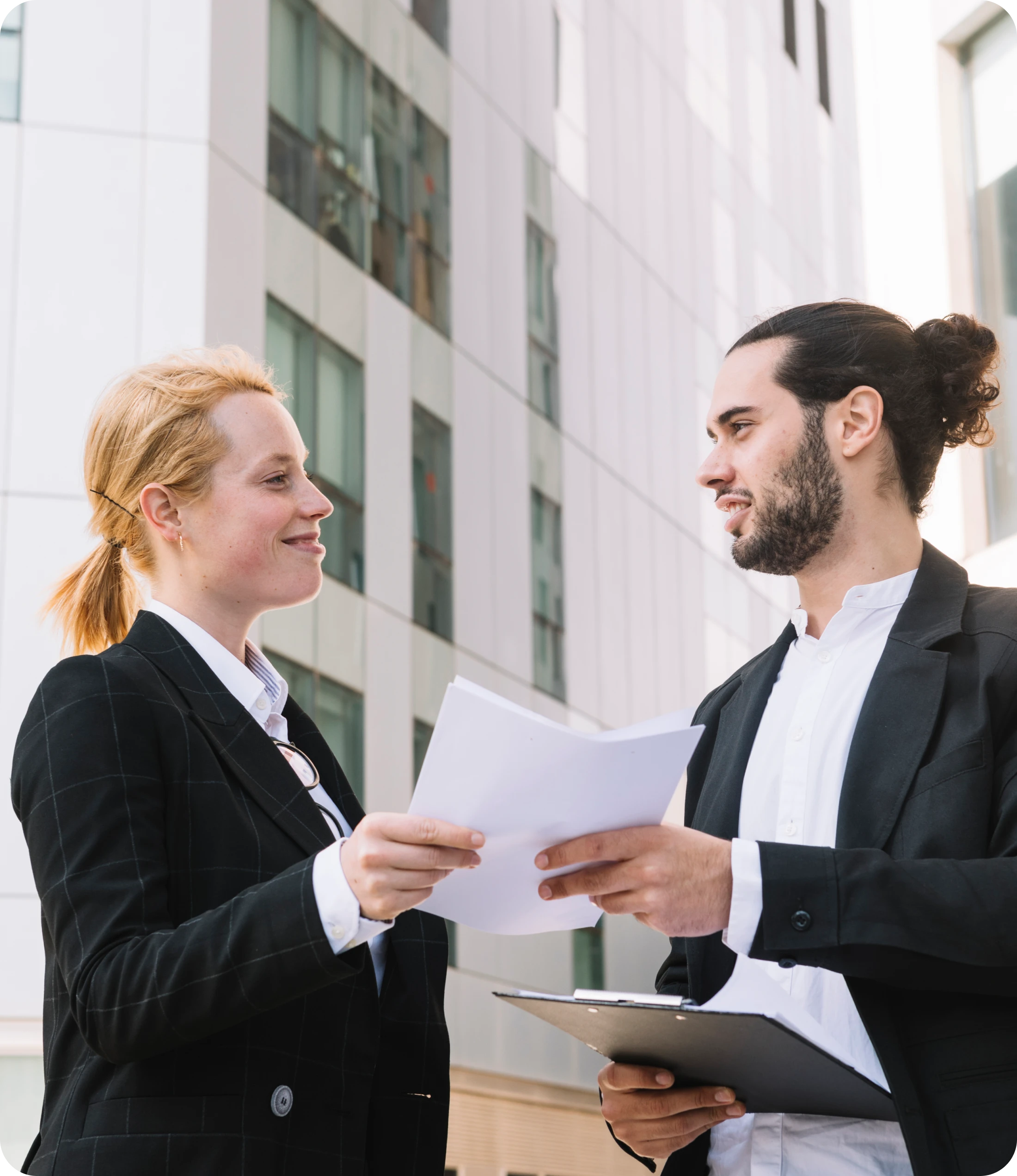 Blonde Woman In Black Blazer & Bearded Man Holding Contract Papers Outside A White Building