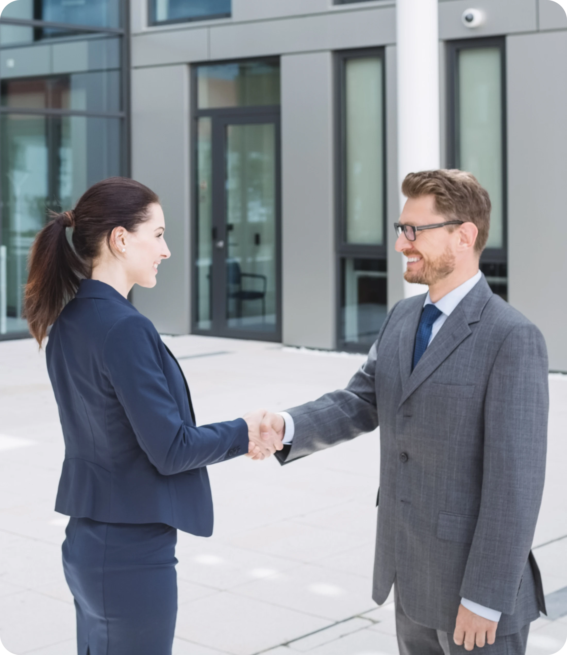 Woman In Navy Suit & Man In Grey Suit Shaking Hands Outside A Glass Office Building