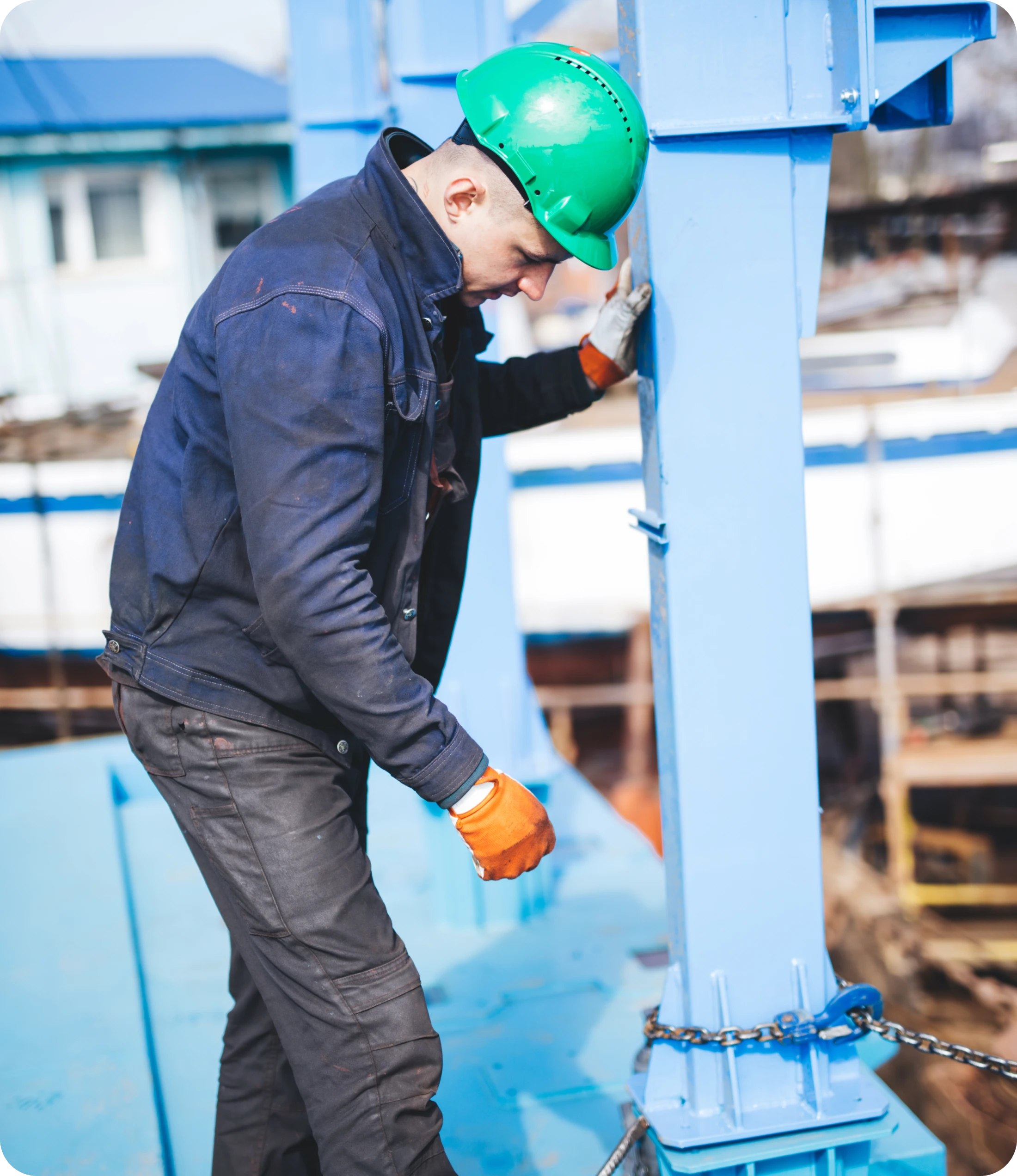 Worker In Green Hard Hat & Orange Gloves Working On A Blue Steel Beam With Chain